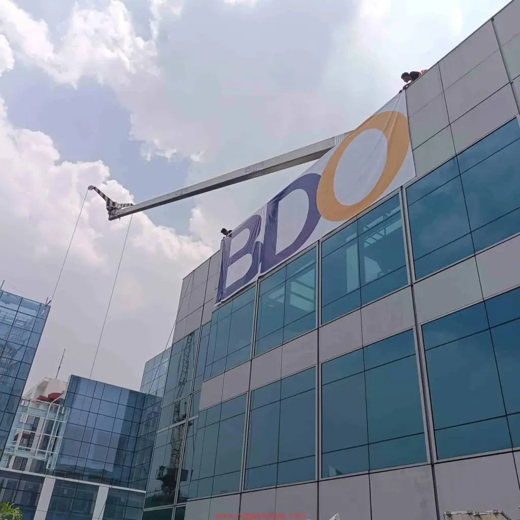 Workers install large 3D acrylic office signage on a modern glass building in Ortigas under clouds, by M&G Global Ads.