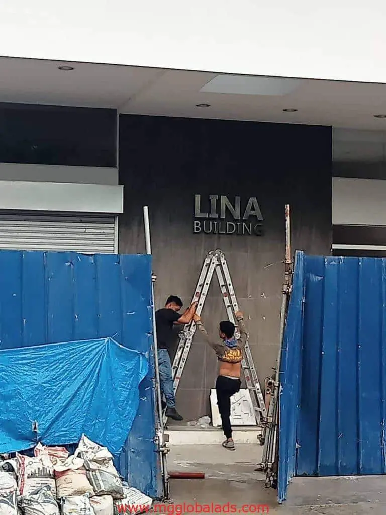 Workers adjusting ladder at LINA BUILDING in Makati, installing new building sign, blue barriers visible by M&G Global Ads.