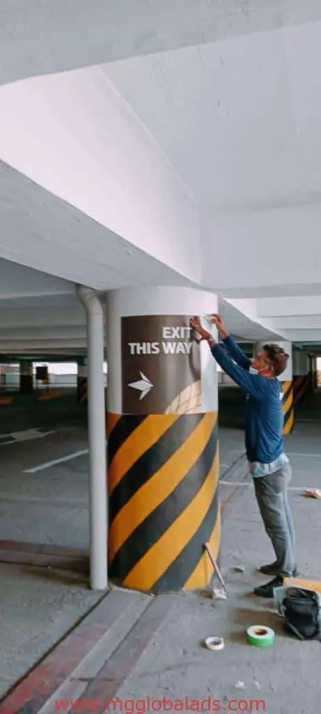 Directional EXIT THIS WAY metal signage installation in Makati parking garage, blue-shirted worker. By M&G Global Ads.
