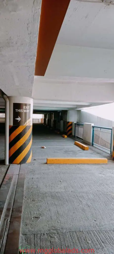 Exit This Way metallic wayfinding sign in Makati parking garage, concrete pillars, yellow stops; sunlight on floor by M&G Global Ads.