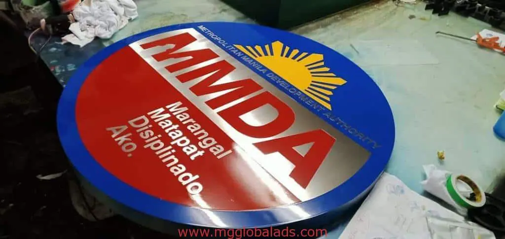 MMDA round stainless engrave building signage for Makati featuring sun emblem and brand values on blue-red, worktable scene by M&G Global Ads.