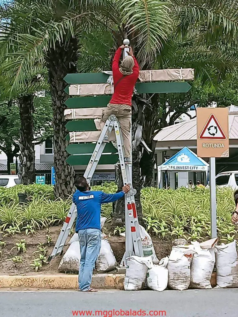 A man on a ladder installing a metallic billboard sign on a street pole in Makati by M&G Global Ads.