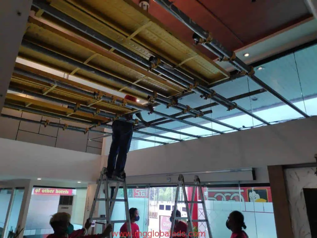 Workers install ceiling panels and pipes near wayfinding signage in a Makati office lobby with glass windows by M&G Global Ads.