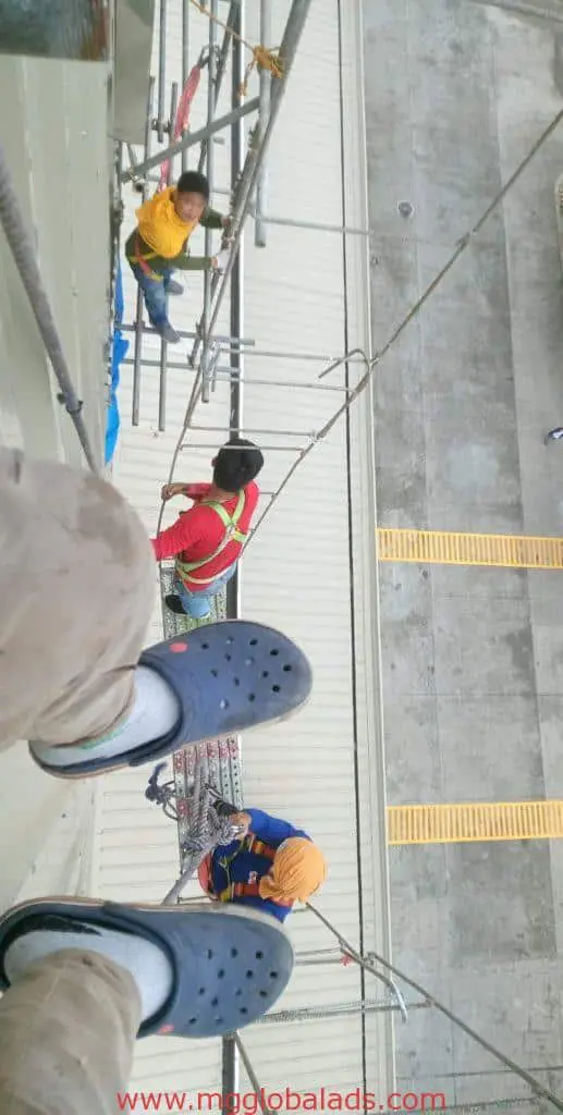 Billboard installation for SM client in Ortigas, workers on ropes and scaffolding; blue clogs visible. By M&G Global Ads.