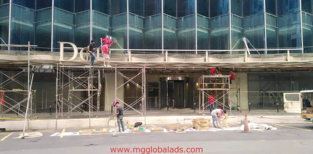 Workers install acrylic building signage for a corporate client in Ortigas, using scaffolding and tools. By M&G Global Ads.