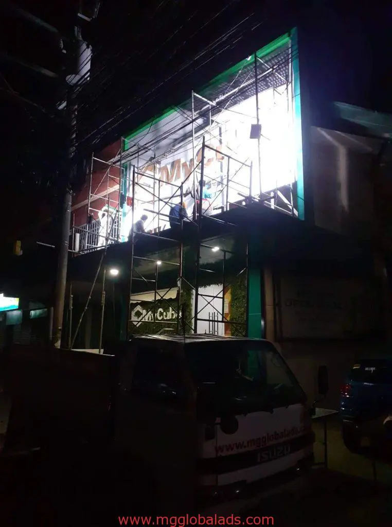 Workers on scaffolding installing LED building signage at night in Makati, truck below, power lines overhead, by M&G Global Ads.