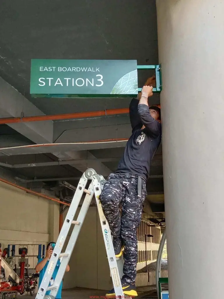Metallic wayfinding signage installation for EAST BOARDWALK STATION 3 in Ortigas by M&G Global Ads. Worker on ladder, covered walkway.