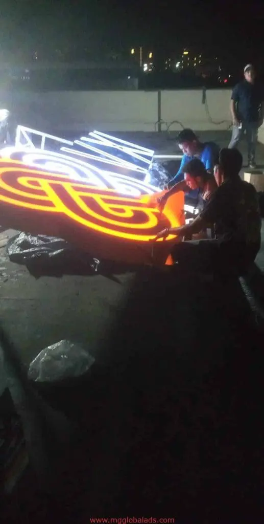 Workers assembling illuminated outdoor signage on a rooftop in Makati at night, glowing orange and white letters by M&G Global Ads.