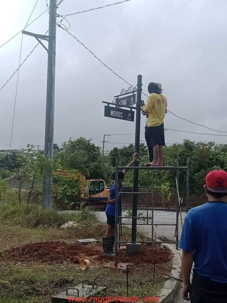 Metal street signage installation labeled WOODS LN in Makati with scaffolding and tools by M&G Global Ads.