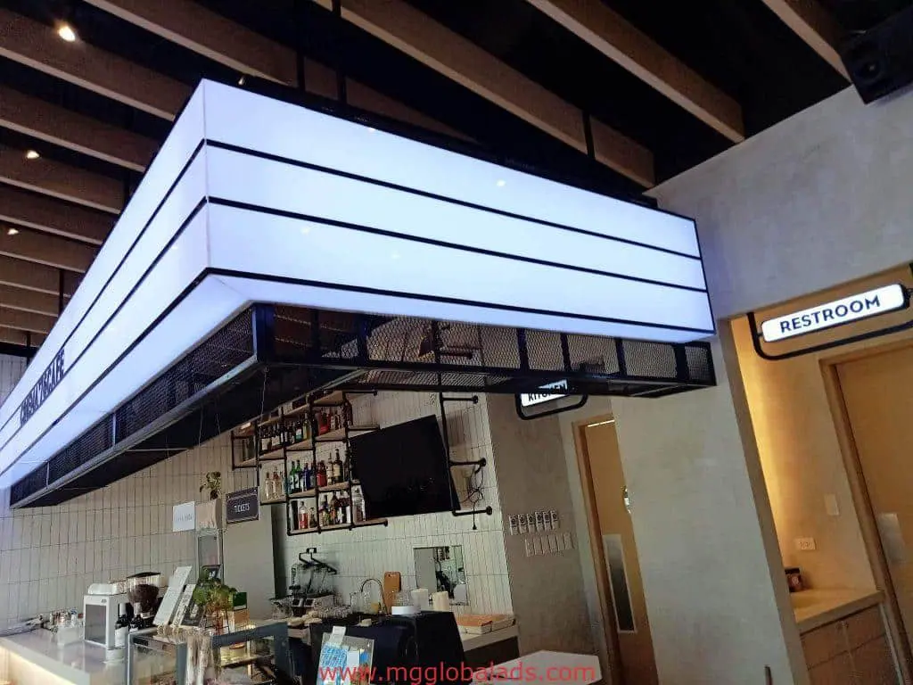 Modern café in Makati with acrylic restroom signage above doorway by M&G Global Ads; counter, bottles, TV, and bright ceiling light.