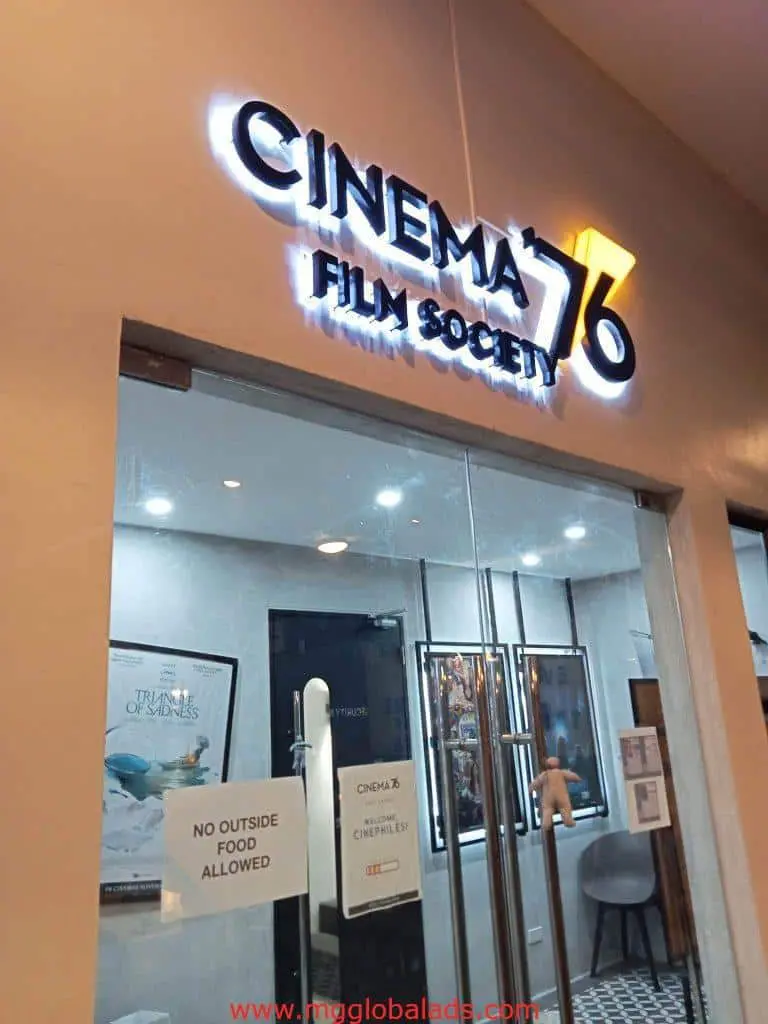 Cinema 76 LED signage at glass entrance in Quezon City with interior posters, chairs, and no outside food sign by M&G Global Ads.