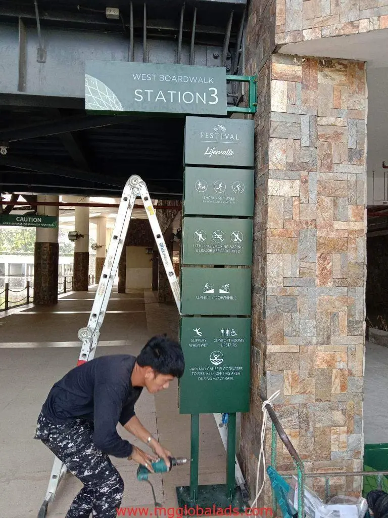 Acrylic rules signage installation at West Boardwalk Station 3, Quezon City by M&G Global Ads. Man drills near ladder, stone wall.
