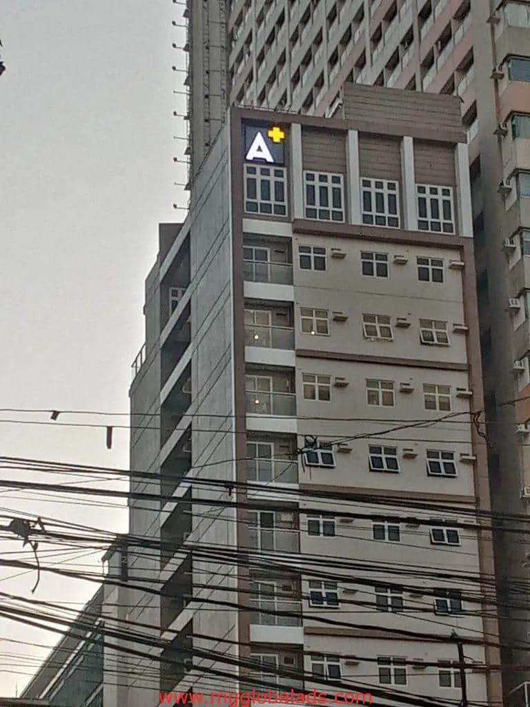 Illuminated metallic building signage with A and yellow plus sign on top of Makati tower, cloudy sky, by M&G Global Ads.