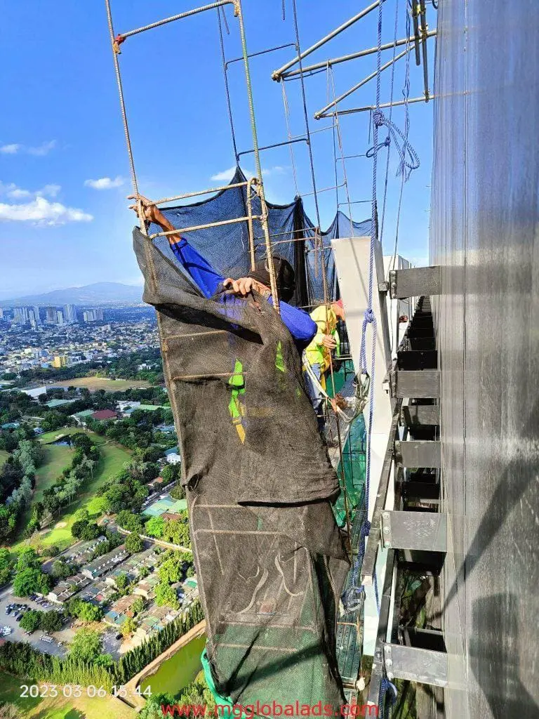 Workers install outdoor building signage and a black safety net on a high-rise in Makati cityscape by M&G Global Ads.