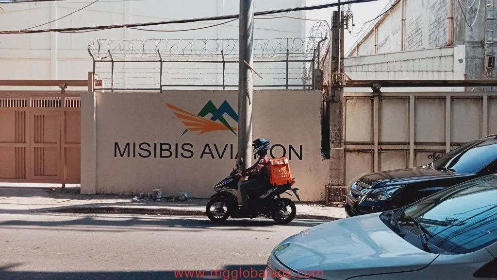 Metallic MISIBIS AVIATION signage and logo on wall in Makati, with delivery rider passing by—by M&G Global Ads.