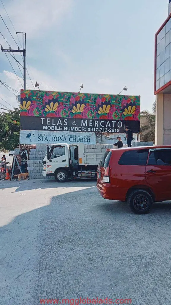 Outdoor floral signage for Telas de Mercado and Sta. Rosa Church in Makati, with truck unloading bricks by M&G Global Ads.