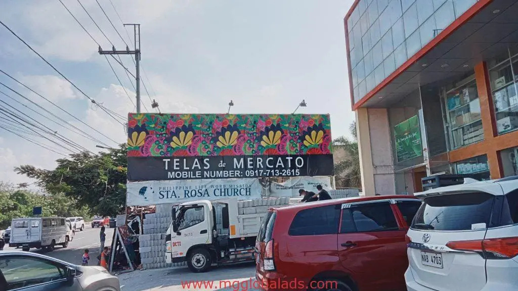 Outdoor building signage for Telas de Mercado with floral designs in a Quezon City parking lot—vehicles and street view—by M&G Global Ads.