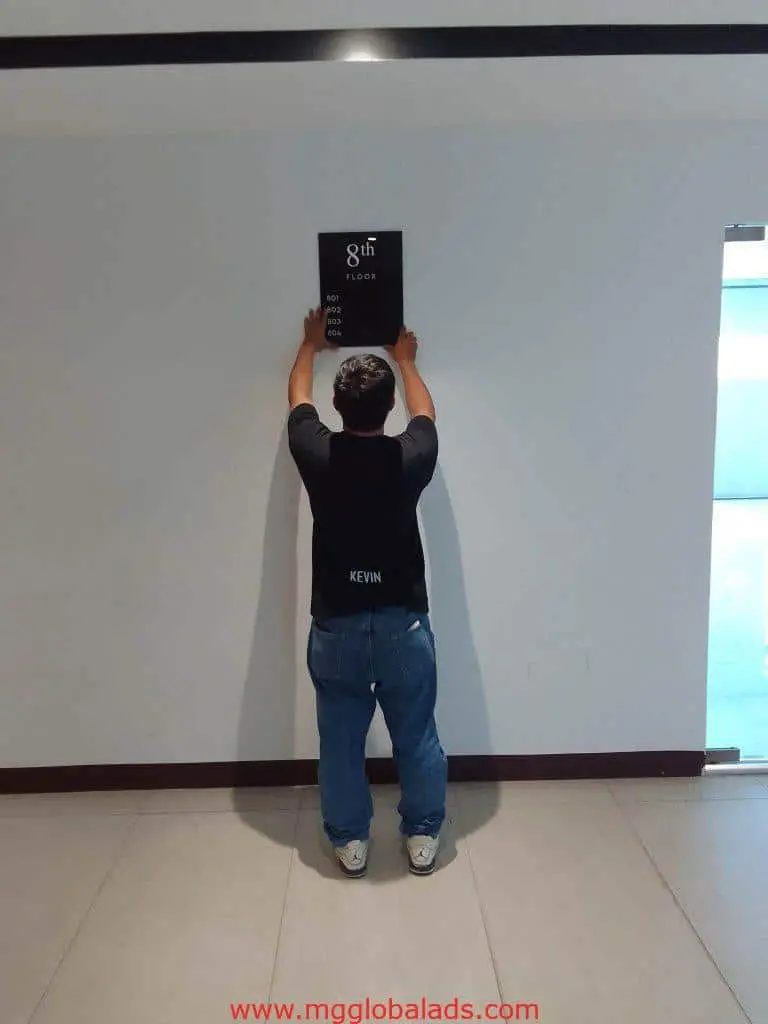 Indoor acrylic 8th Floor directional signage installation in Makati, by M&G Global Ads. Person adjusting sign with KEVIN shirt.