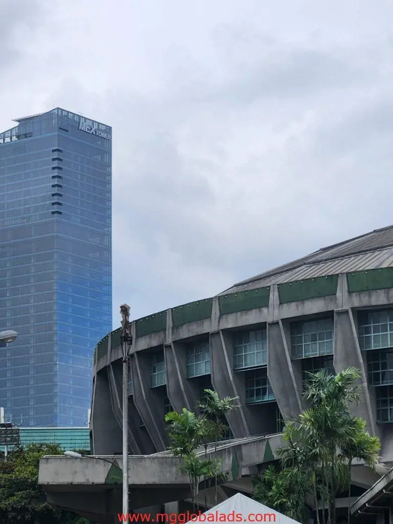 Outdoor billboard signage in front of city buildings and palm trees in Makati, cloudy sky overhead, by M&G Global Ads.