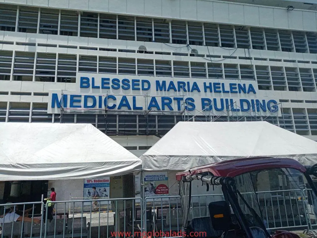 Medical Arts Building blue outdoor signage for Blessed Maria Helena in Quezon City, with tents and red vehicle, by M&G Global Ads.