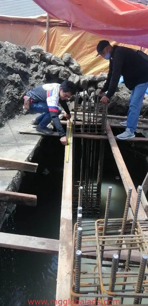 Two workers measure a concrete foundation near billboard signage in Makati, with steel rebar and planks. By M&G Global Ads.