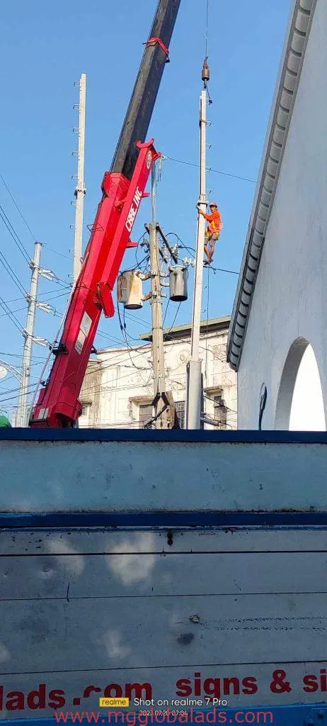 Worker in safety gear repairing electrical wires near a white building with storefront signage in Makati by M&G Global Ads.