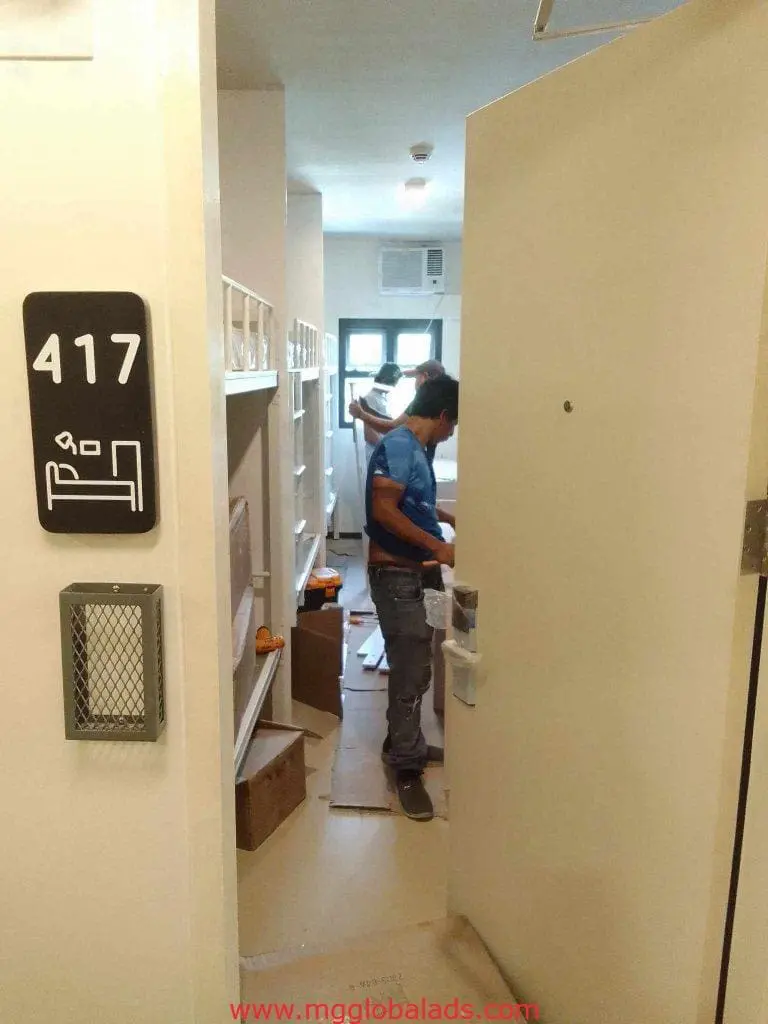 Metallic dorm room signage 417 in Makati, bunk beds and boxes visible, people organizing. By M&G Global Ads.