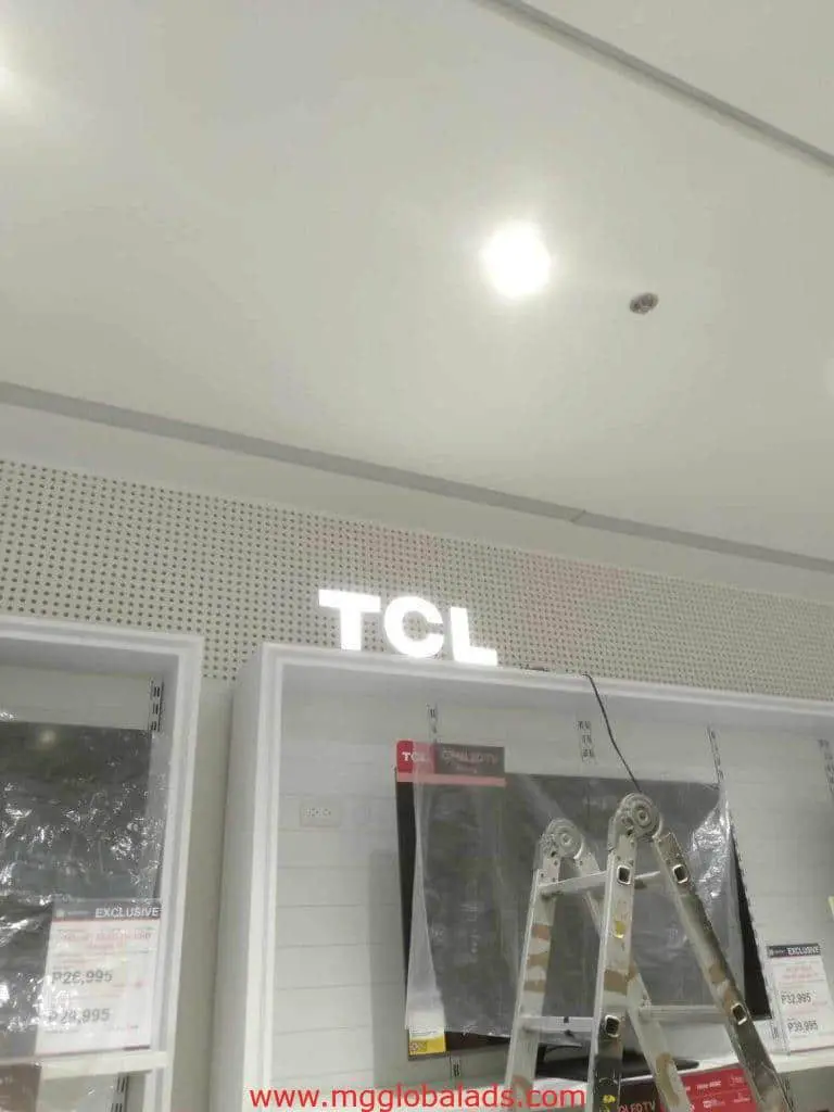 Metallic TCL indoor store signage on a white wall in Quezon City with step ladder and shelves, by M&G Global Ads.