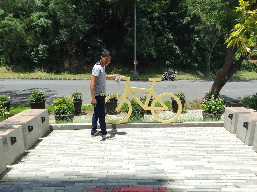 A person beside a large yellow bicycle-shaped outdoor sculpture signage in Makati, greenery and road visible, by M&G Global Ads.