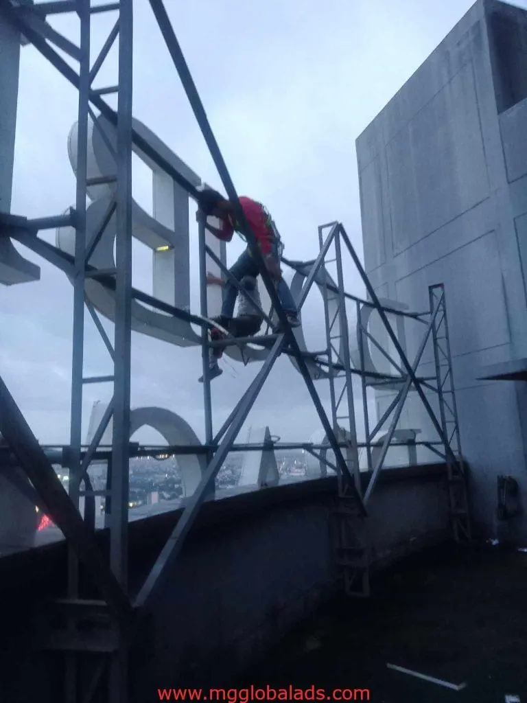 Worker installing large LED billboard signage on rooftop at dusk in Ortigas, cityscape behind, by M&G Global Ads.