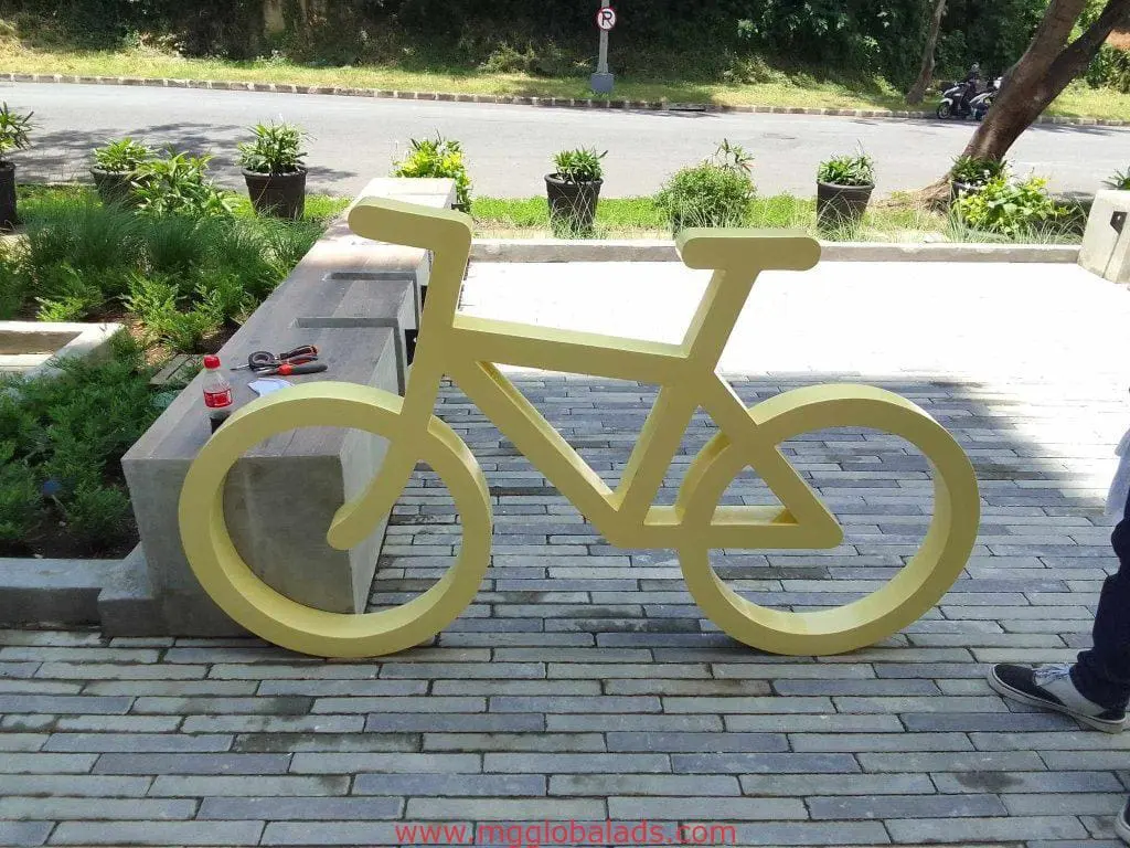 Yellow bicycle-shaped bike rack with building signage near sidewalk in Makati, tools on bench, motorcycles parked—by M&G Global Ads.