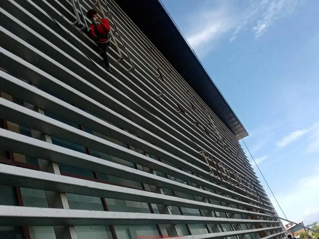 A worker on ropes cleans glass near building signage in Makati with metal beams and blue sky, by M&G Global Ads.