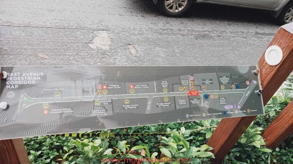 Outdoor pedestrian corridor signage map on glass panel in Quezon City with labeled icons, car, and greenery by M&G Global Ads.