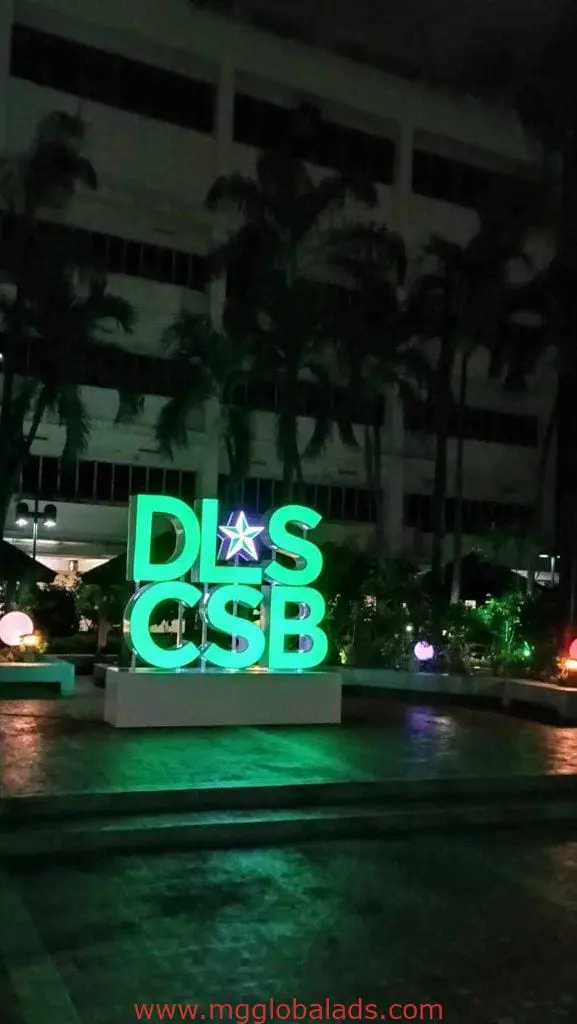 Outdoor green LED signage for DLS CSB at night in Makati, surrounded by palm trees and colorful lights, by M&G Global Ads.