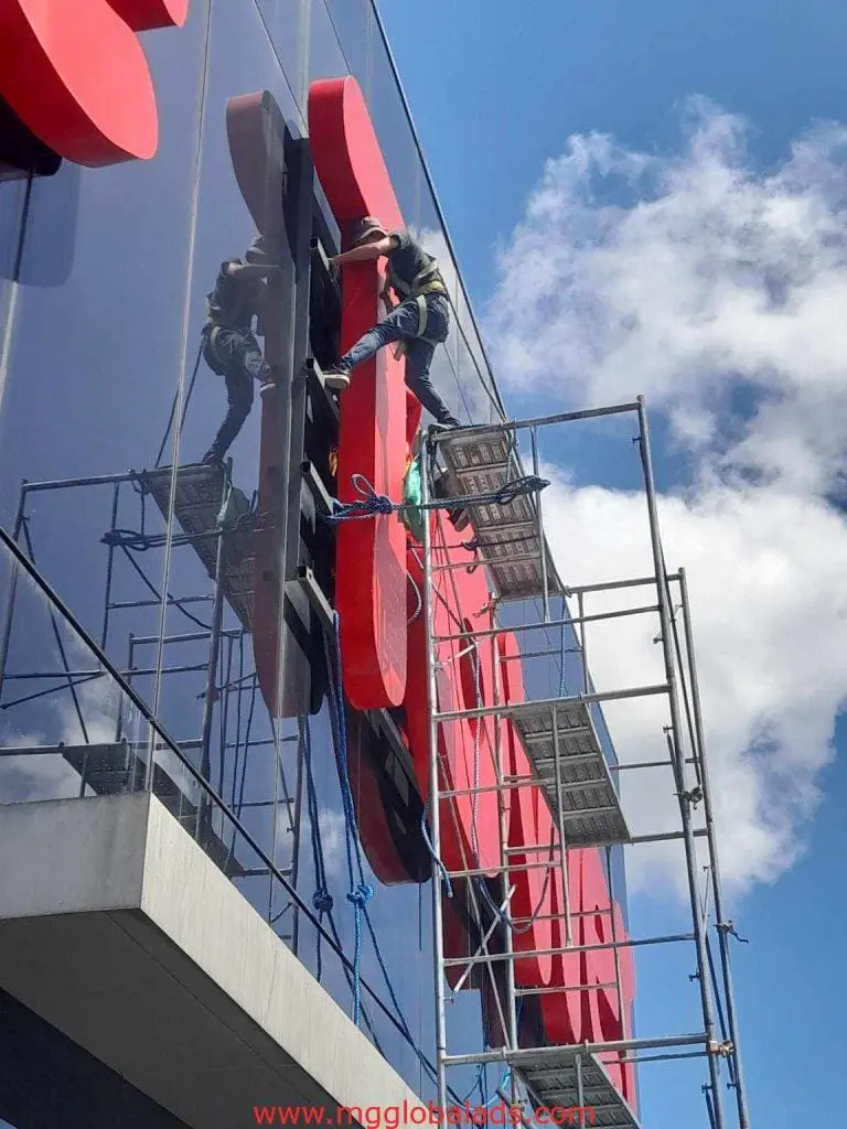 LED signage installation by worker on scaffolding in Makati, red LED sign on building with glass facade by M&G Global Ads.