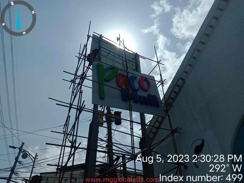 Acrylic Paco Mart signage installation by workers on scaffolding in Quezon City under a cloudy sky by M&G Global Ads.