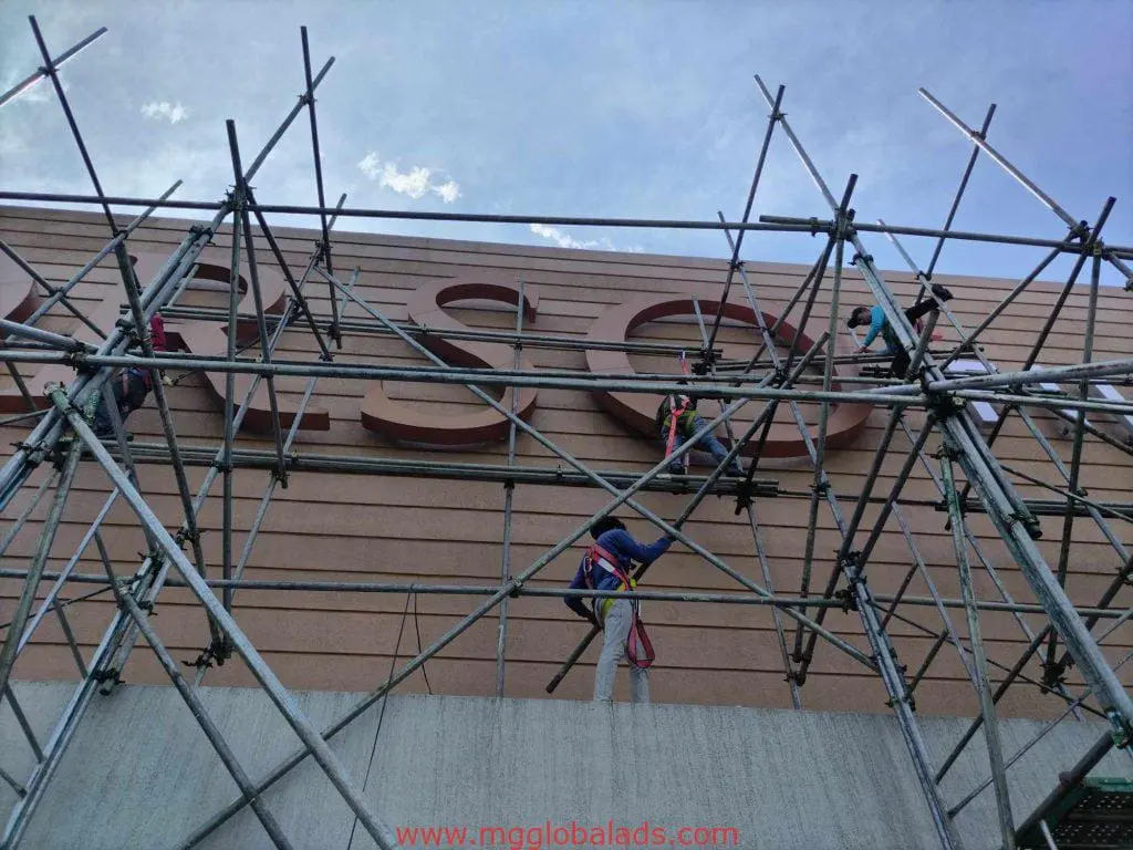 Workers install bold letter outdoor signage on scaffolding at a large facade in Makati, partly cloudy sky, by M&G Global Ads.