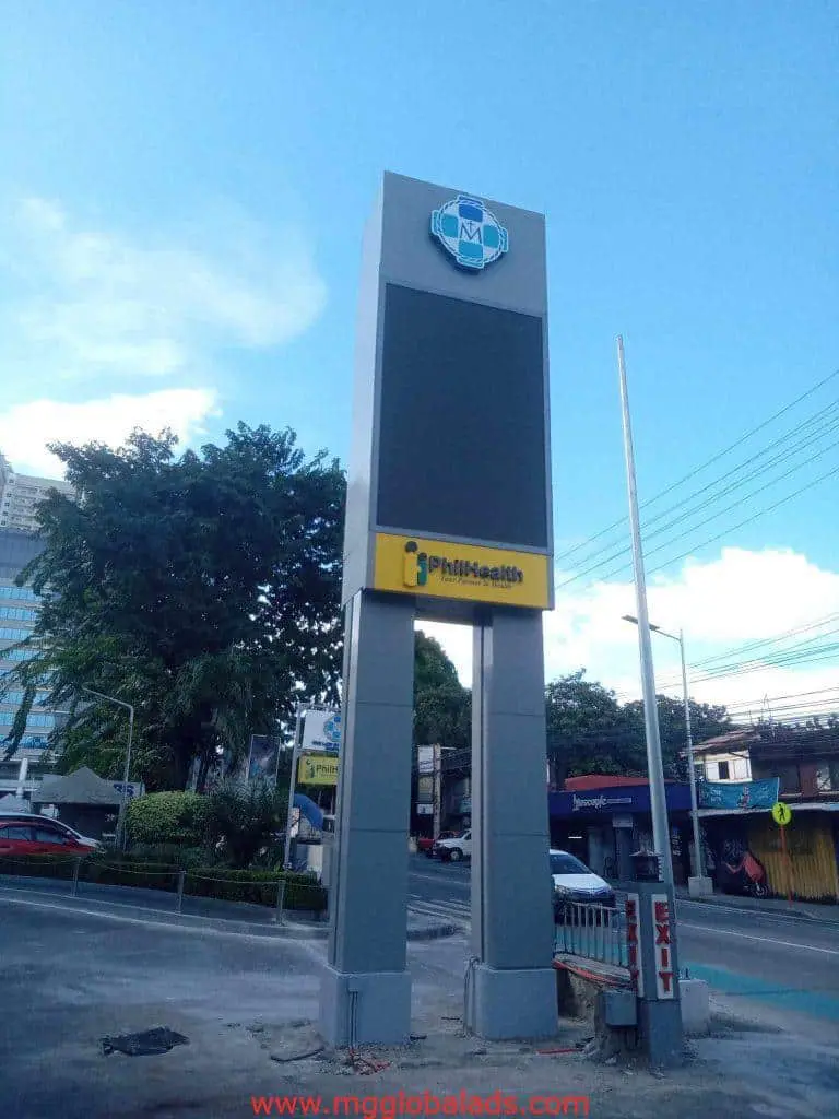 PhilHealth pylon signage with NM logo in Quezon City, surrounded by trees, buildings, and power lines by M&G Global Ads.