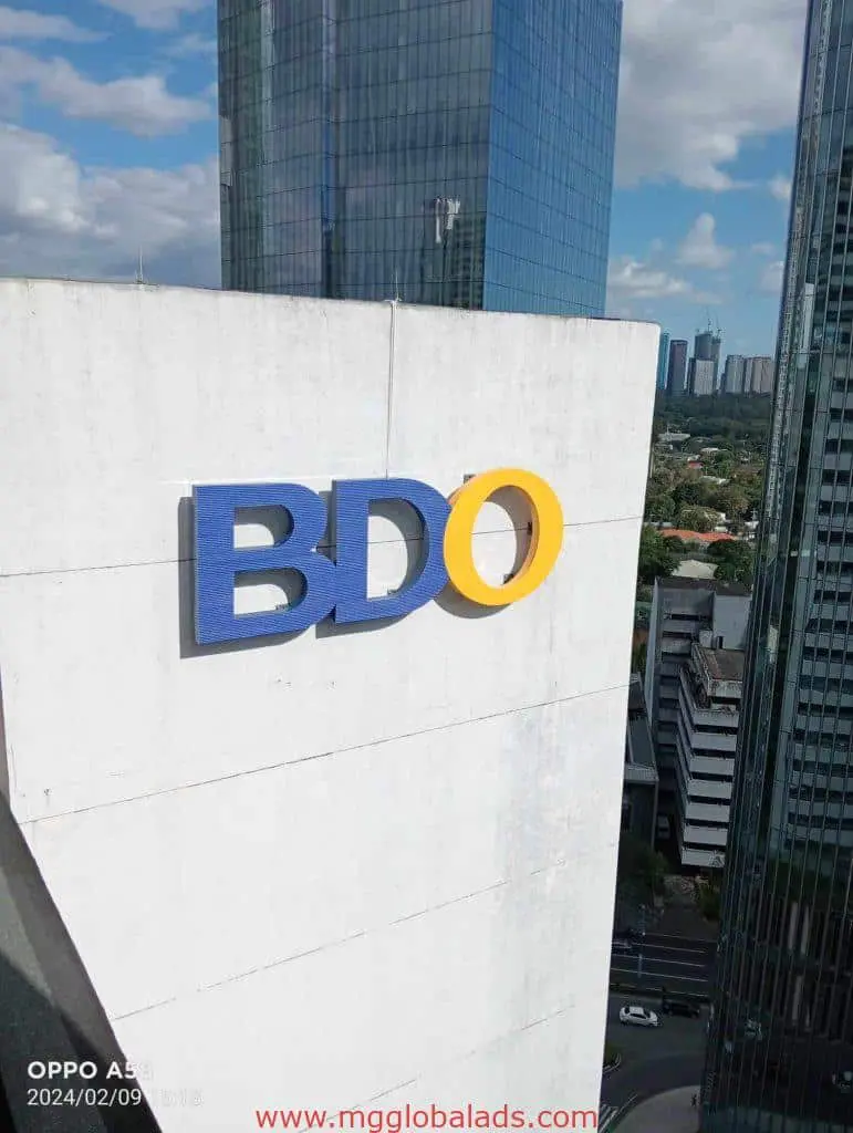 BDO outdoor billboard signage on white building in Makati, city skyscrapers and trees in background, by M&G Global Ads.