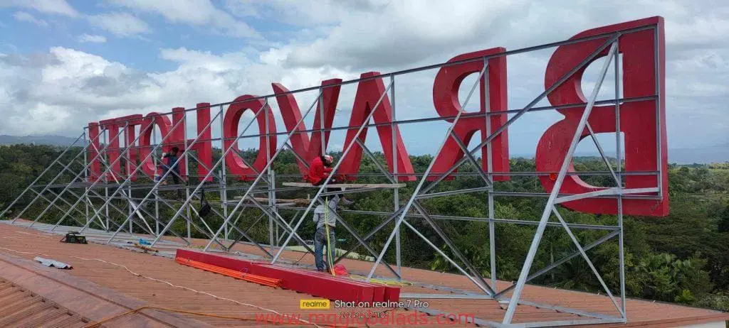 Makati rooftop LED signage installation with workers handling bold red letters, cloudy sky and trees, by M&G Global Ads.