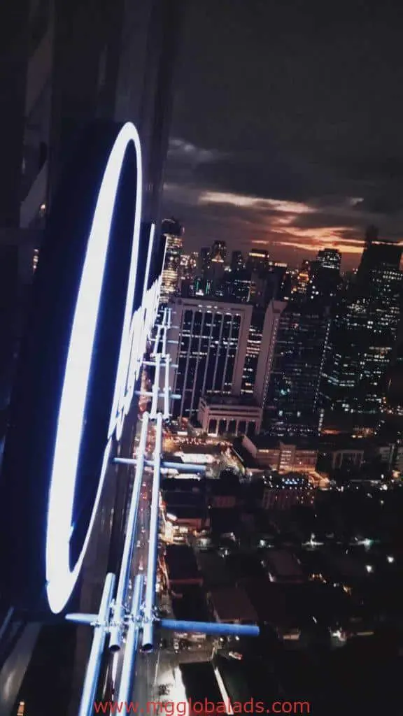 Neon metallic building signage at dusk in Makati, city skyscrapers and streets glowing under clouds by M&G Global Ads.