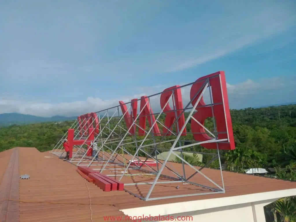 Backlit outdoor signage with large red PRINCE letters on a metal frame in Quezon City, rooftops and trees by M&G Global Ads.