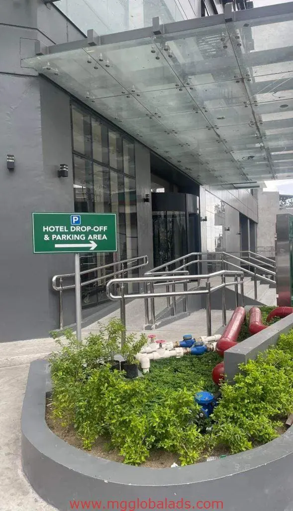 Green metallic hotel drop-off & parking area sign at entrance in Makati, with glass canopy and plants by M&G Global Ads.
