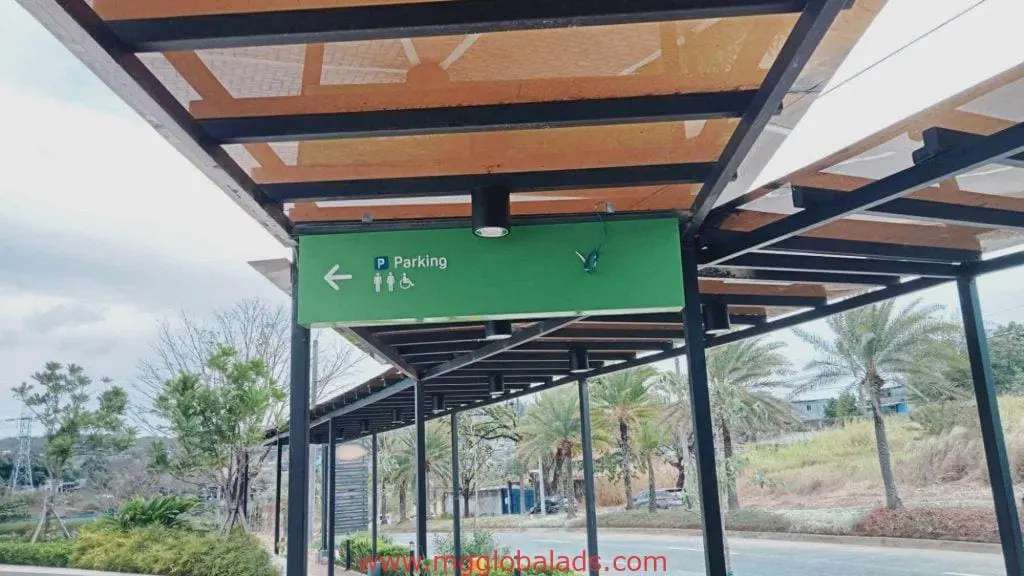 Green acrylic directional signage for parking, restrooms, accessible access in Makati by M&G Global Ads. Palms and road behind.