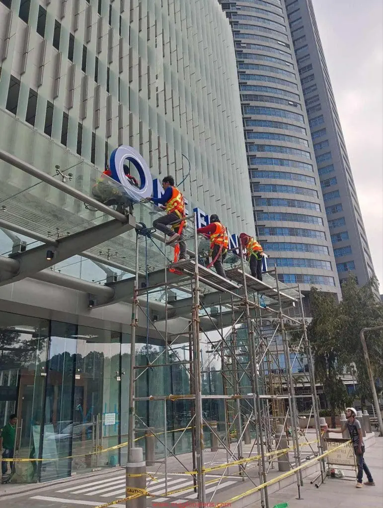 Workers install acrylic building signage for a corporate client in Makati on a glass canopy, with skyscraper view, by M&G Global Ads.
