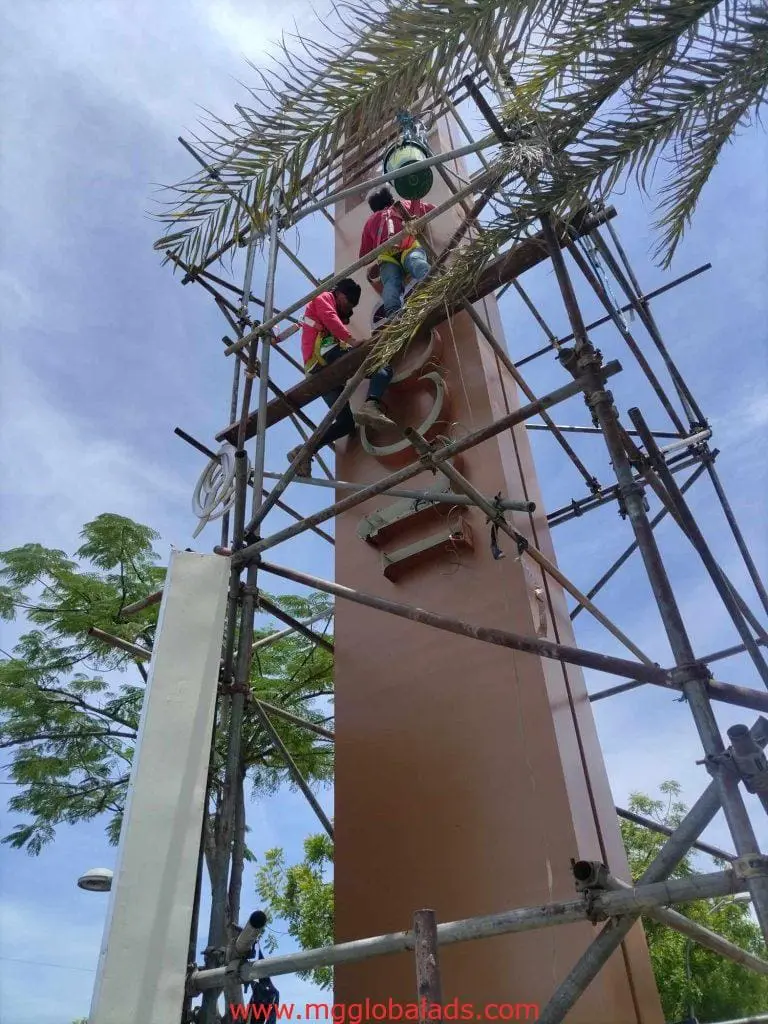 Workers on scaffolding install building signage in Ortigas, both wearing safety gear under clear sky. By M&G Global Ads.