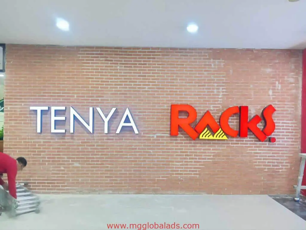 LED signage for Tenya and Racks on a brick wall, installation in Makati by M&G Global Ads. A worker adjusts a ladder nearby.