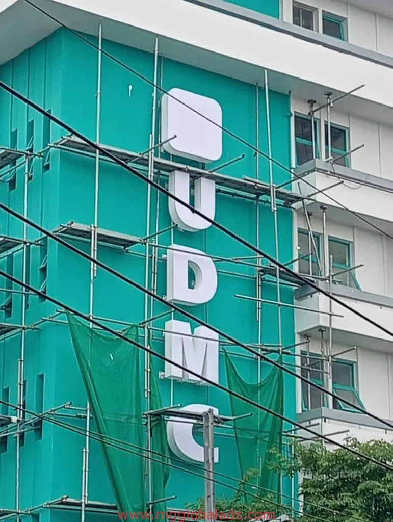 Vertical UDMC building signage on tall teal and white site in Ortigas with scaffolding and wires, by M&G Global Ads.