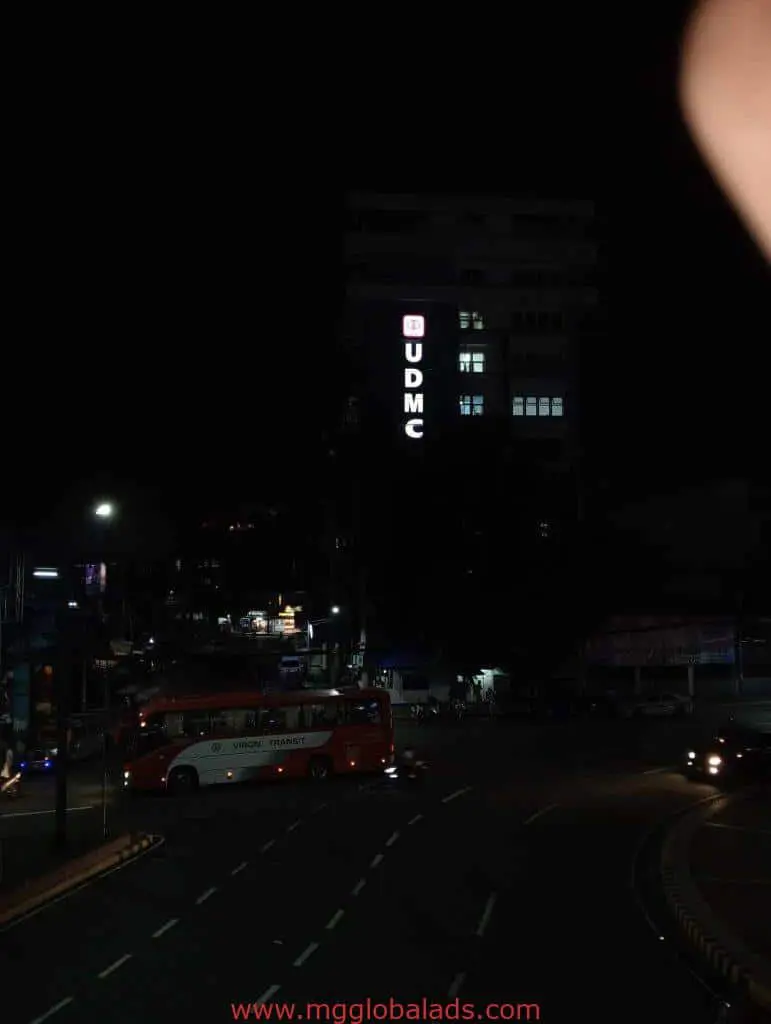 Vertical LED signage for UDMC on a tall building in Quezon City at night, red and white bus in front. By M&G Global Ads.