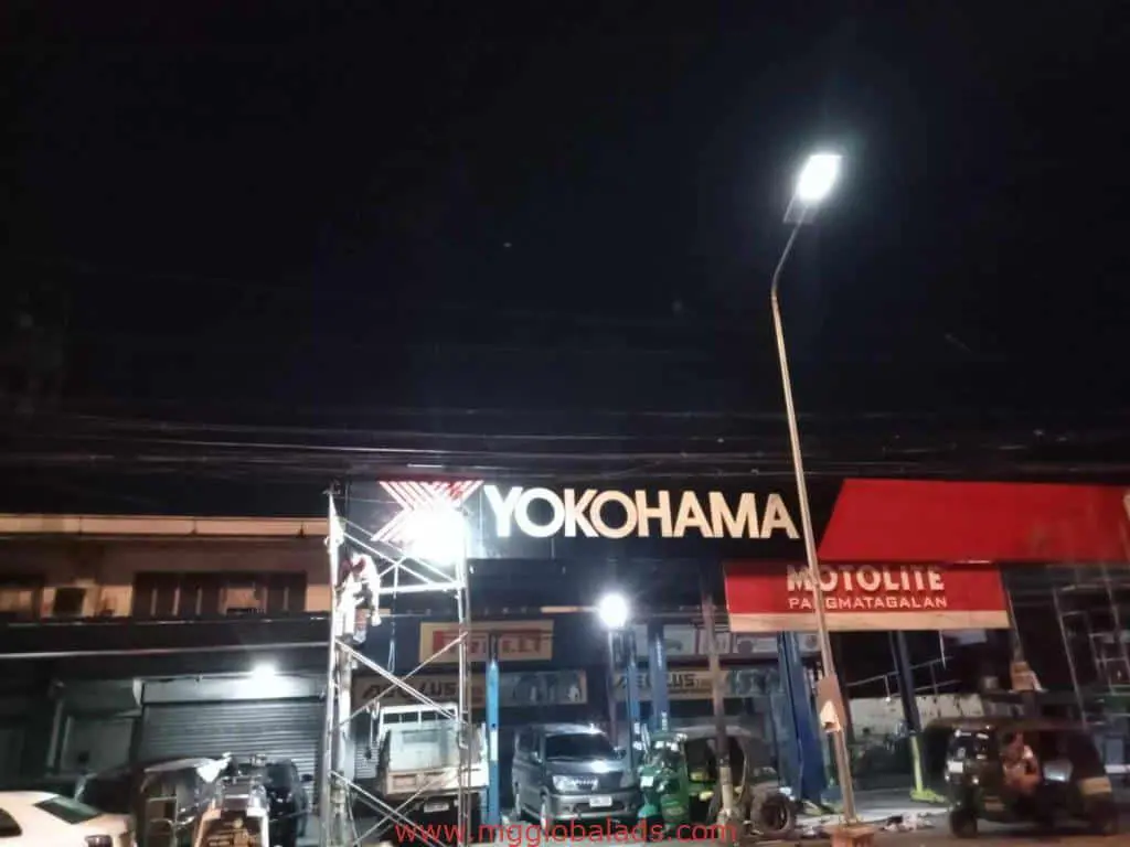 Yokohama acrylic signage in Makati at night, with scaffolding, workers, vehicles, and Motolite sign by M&G Global Ads.